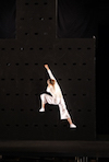 Detail from a photo of a metacreated dance performance. A dancer in loose white clothes performs against a black background.